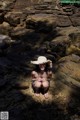 A woman in a straw hat sitting on a rocky beach.