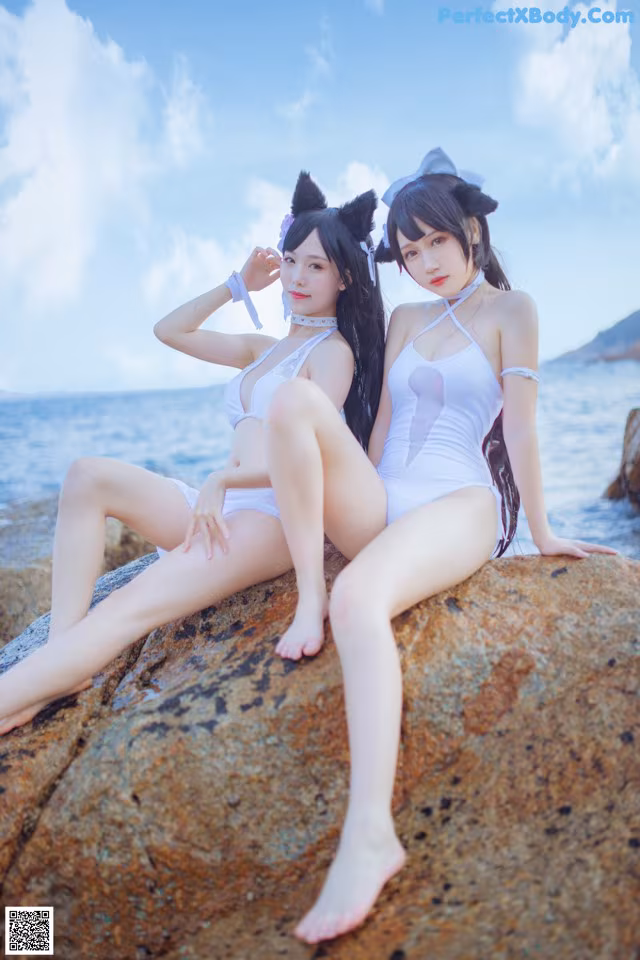 Two women in white bathing suits sitting on a rock by the ocean.